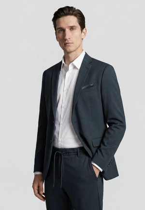 Mann trägt dunkle Anzugjacke, weißes Hemd und dunkle Hosen mit Kordelzug, steht vor hellem, einfachem Hintergrund.