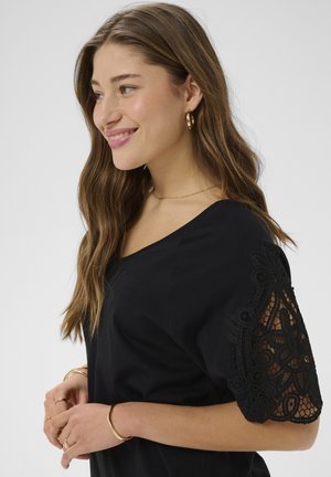 Fiatal nő hosszú hullámos hajjal, arany karika fülbevalóval, fekete felsővel csipkés ujjakkal és arany karkötőkkel, mosolyog és balra néz.