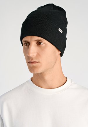 Cappello nero a maglia con texture a coste, design aderente e una piccola etichetta bianca. Indossato con una maglietta bianca semplice con scollo rotondo.