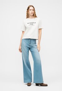 Γυναίκα με μακριά κυματιστά μαλλιά που φοράει λευκό τυπωμένο μπλουζάκι, ανοιχτό μπλε τζιν παντελόνι με φαρδιά μπατζάκια και καφέ μποτάκια με τακούνι, όρθια σε λευκό φόντο.