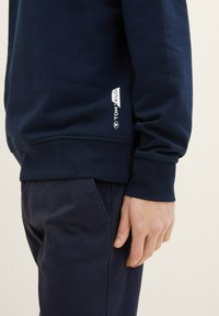 Granatowy sweatshirt z długimi rękawami, ściągaczami przy rękawach i dolnej krawędzi, z małą białą metką z logo po boku. Wykonany z miękkiego materiału z bawełny.