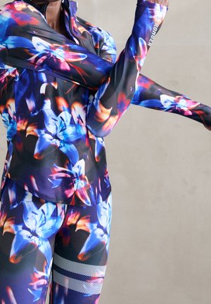 Persoon die de armen strekt, gekleed in levendige sportkleding met een bloemmotief in blauw, paars en rood tegen een effen achtergrond.