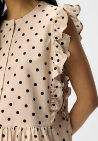 Blouse rose clair avec des pois noirs, présentant des manches volantes et un devant à boutons. Tissu lisse avec une taille froncée.