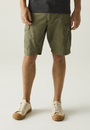 Uomo con pantaloncini cargo verde oliva, sneakers beige con suola marrone e una maglia scura, in piedi su uno sfondo chiaro semplice.