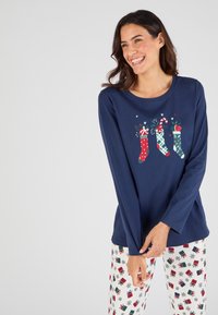 Camiseta de manga larga en color marino con un gráfico de tres medias coloridas; combinada con pantalones blancos que presentan un patrón festivo de regalos en rojo y verde.
