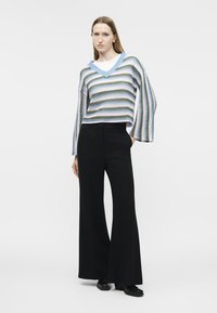 Femme portant un pull à rayures bleues, blanches et vertes avec un col en V, un pantalon large noir et des chaussures noires, debout devant un fond blanc.