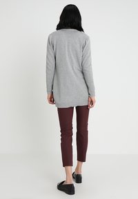 Kvinna med svart hår som bär en lång grå tröja, vinröda byxor och svarta loafers, stående mot en enfärgad ljus bakgrund.