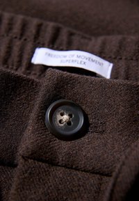 Brązowa tkanina o miękkiej fakturze z czarnym guzikiem z czterema otworami. Metka z napisem "WOLNOŚĆ RUCHU SUPERFLEX". Zdjęcie z bliska.