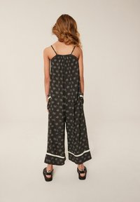 Salopetă neagră cu bretele subțiri, model floral, pantaloni largi, accente din dantelă la fund și buzunare laterale. Modelul stă cu spatele la cameră.