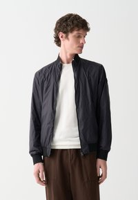 Jachetă bomber ușoară, neagră, cu fermoar frontal, manșete ribbate și guler prevăzut cu un design. Purtați-o peste un pulover alb și pantaloni maro.