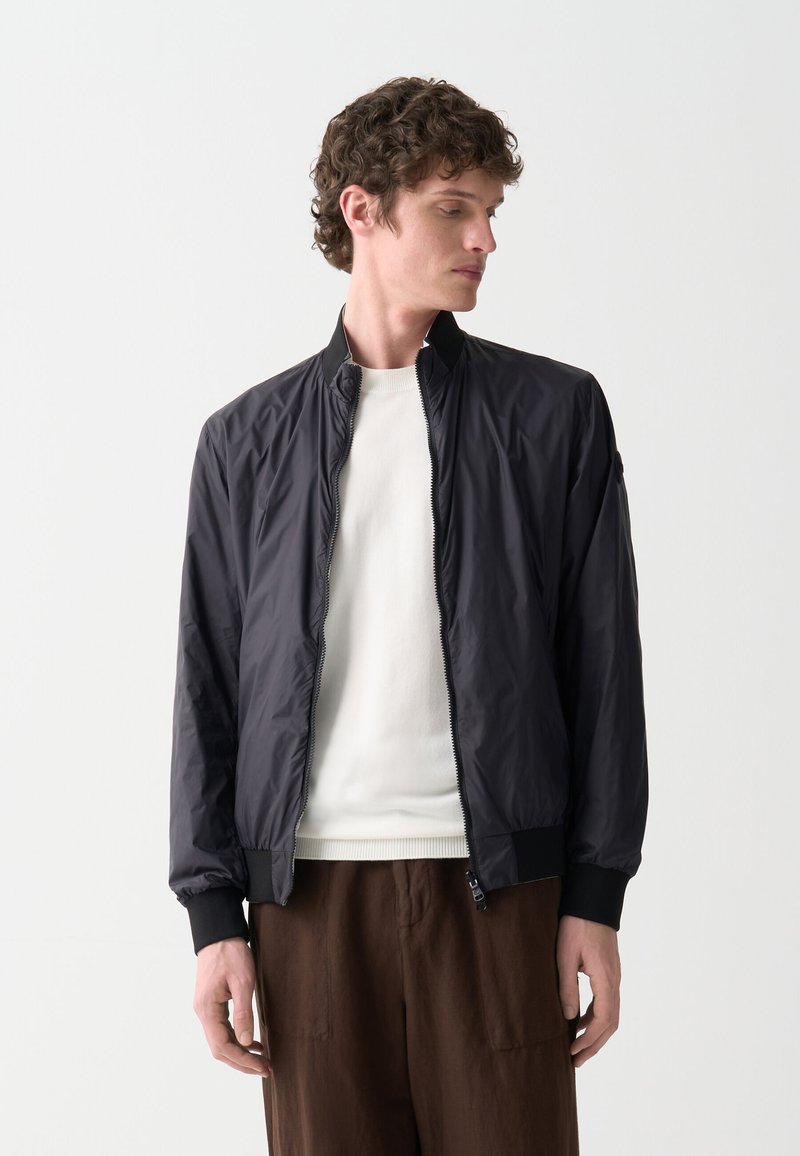 Jachetă bomber ușoară, neagră, cu fermoar frontal, manșete ribbate și guler prevăzut cu un design. Purtați-o peste un pulover alb și pantaloni maro.