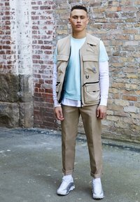 Jeune homme debout contre un mur en briques, portant un gilet cargo beige, une chemise bleu clair sur des manches longues blanches, un pantalon beige et des baskets blanches.
