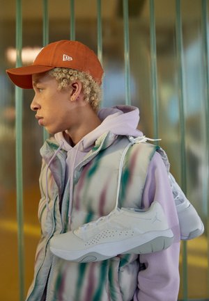 Junge Person mit lockigem blondem Haar, die eine orangefarbene Kappe und eine Batikweste trägt, trägt weiße Sneakers über der Schulter vor vertikalen Stangen.