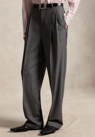 Person trägt hoch taillierte, weit geschnittene graue Hose mit vorderen Falten, schwarzen Gürtel mit silberner Schnalle, schwarze spitze Schuhe und hellrosa Hemd.