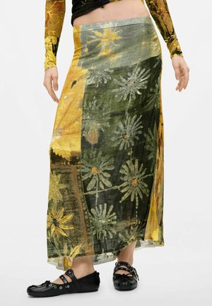 Person iført en lang nederdel med gult og grønt blomsterprint, sort top og sorte sko med spænder står på en ensfarvet baggrund.