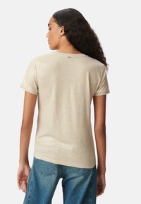 Vrouw met lang krullend haar, gekleed in een beige shirt met korte mouwen en blauwe denimjeans, gezien van achteren tegen een witte achtergrond.