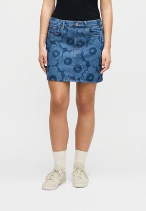 Kvinde iført en blå denim mini nederdel med store blomsterprint, hvide sokker, beige sneakers og en sort top, synlig fra taljen og ned.