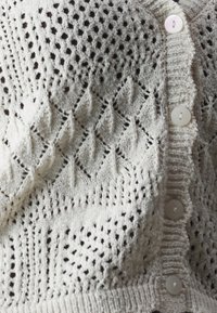 Ljust beige stickad kofta med ett strukturerat, öppet mönster, rundad halsringning och fem vita knappar fram.