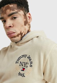 Felpa beige con logo ricamato "Tommy Jeans NYC" e colori accento, caratterizzata da un tessuto texturizzato e una tasca a marsupio frontale.