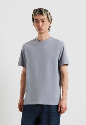 Giovane uomo con capelli corti biondi e scuri che indossa una maglietta grigia chiara tinta unita a maniche corte e jeans blu scuro su sfondo bianco.