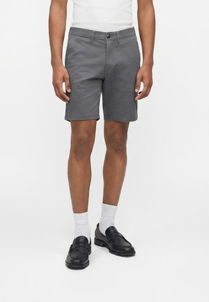 Uomo che indossa pantaloncini grigi su misura, calzini bianchi, mocassini neri e una camicia bianca su uno sfondo chiaro e uniforme.