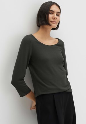 Femme aux cheveux courts noirs, portant un haut à manches longues gris foncé et un pantalon noir, appuyée contre un mur blanc, les mains dans le dos.
