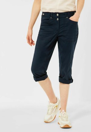 Persoon draagt opgerolde donkerblauwe capribroek met voorzakken en witte sneakers met beige en gele accenten.