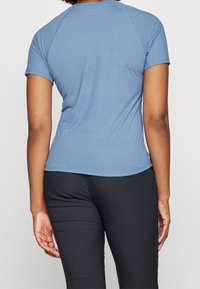 Modrá košile s krátkými rukávy z měkké látky, s kulatým výstřihem a raglánovými rukávy. Nasažená s černými kalhotami, ukazující zaoblený lem.