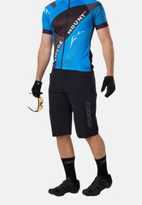 Cycliste masculin portant un maillot à manches courtes bleu et noir et un short noir, tenant des lunettes de sport teintées jaunes et portant des gants noirs.