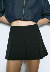 Schwarzer, plissierter Minirock mit glatter Textur und strukturiertem Taillenbund; kombiniert mit einer kurzen Jacke aus dunklem Denim.
