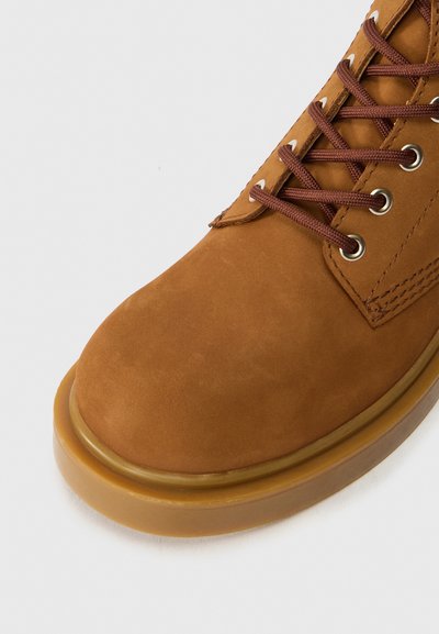 Bota de tobillo de ante marrón con punta redondeada, cordones marrones, ojales en tono dorado y suela de goma suave color beige.