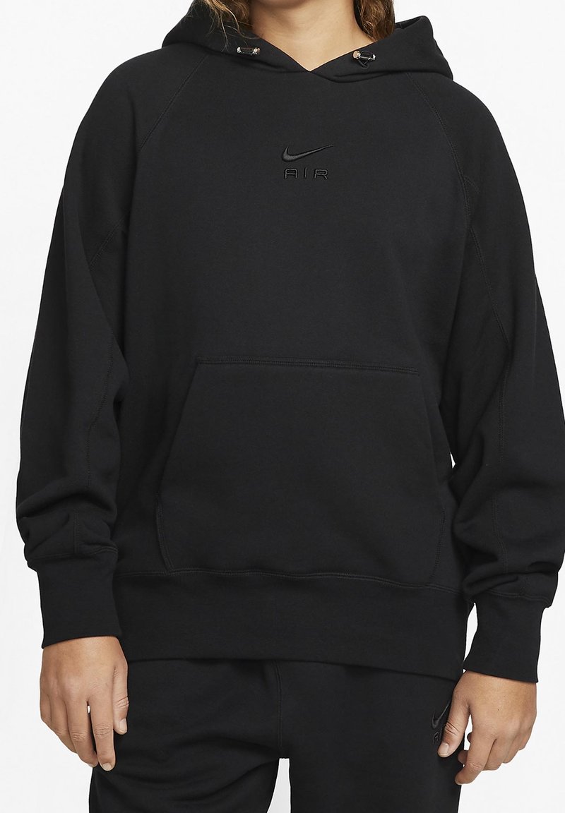 Personne portant un sweat à capuche Nike Air noir et un pantalon noir assorti, debout avec les mains détendues le long du corps, fond blanc.
