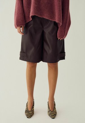 Personne portant un pull rose doux, un short noir jusqu'aux genoux et des talons aiguilles pointus imprimés zèbre, debout sur un fond clair uni.