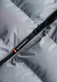Sezione di una giacca a vento grigia con una cerniera nera e un'etichetta. Il tessuto appare liscio con una texture lucida e morbida.