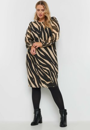 Robe à manches longues et longueur genou en tissu transparent avec un imprimé zèbre noir et beige, présentant des manches bouffantes et une coupe décontractée. Portée avec des bottes noires à la cheville.