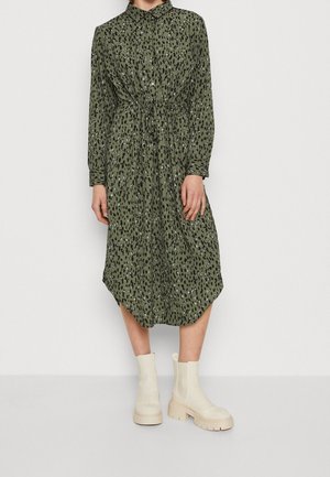 Zelené vzorované šaty na knoflíky se zavazováním v pase, s dlouhými rukávy a předním krátkým, zadním delším okrajem. Kombinováno s krémovými kotníčkovými botami.