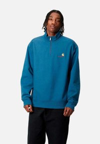 Sudadera de forro polar azul con media cremallera, puños acanalados y un pequeño logo en el pecho. Presenta un ajuste clásico y relajado, con una textura suave.