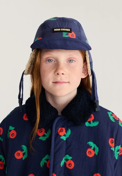 Enfant aux cheveux roux et taches de rousseur portant un chapeau et un manteau bleu marine avec un motif floral rouge et vert, chapeau étiqueté Bobo Choses, regardant vers l'avant.