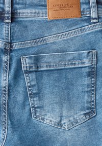 Vaaleat siniset farkut takataskulla, jossa näkyy ompelu ja nahkainen etiketti, jossa lukee "STREET ONE DENIM ATELIER."