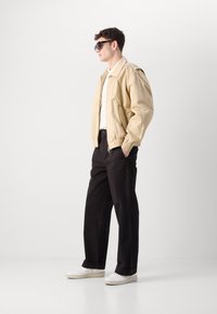 Beige bomber dzseki rugalmas mandzsettával, fehér póló, fekete nadrág és fehér sportcipő. A modell oldalról áll egy világos háttér előtt.