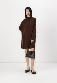 Brun oversized strikkekjole med høy hals, kombinert med en svart lærveske og svarte hæler med dekorativ sløyfedetalj.