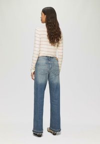 Blaue weit geschnittene Jeans aus Denim mit fransigen Säumen, getragen mit einem gestreiften, hellen Langarmoberteil. Verfügt über zwei Gesäßtaschen und eine entspannte Passform.