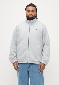 Jachetă gri cu fermoar și guler înalt, cu buzunare laterale. Materialul pare moale și texturat. Purtată cu blugi albastru deschis.