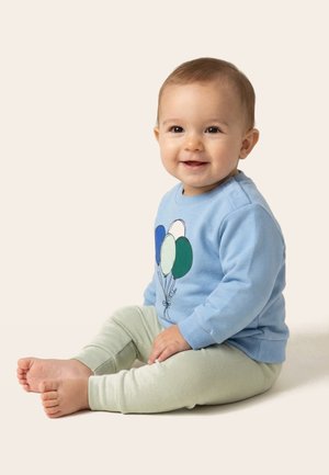 Mosolygós baba ül a padlón, kék pulóvert visel, melyen léggömbös minták vannak, és világos színű nadrágot egy egyszerű háttér előtt.