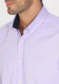 Chemise cintrée violet clair avec un col bleu marine contrastant, un tissu texturé et des boutons blancs le long de la patte de boutonnage.