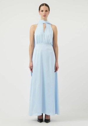 Femme portant une robe longue bleu clair sans manches à col halter avec un décolleté en forme de serrure et des talons noirs pointus.