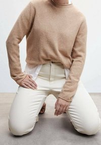 Personne agenouillée sur le sol portant un pull beige par-dessus une chemise blanche et un pantalon taille haute blanc cassé avec des détails de boutons et de coutures visibles.