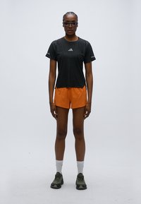T-shirt de sport rayé noir, short orange, chaussures de course gris-vert et chaussettes blanches. Le modèle porte des lunettes et se tient devant un fond uni.