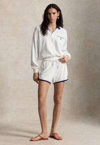 Γυναίκα που φοράει λευκό πόλο με μακρύ μανίκι και ταιριαστό σορτς με ναυτικό γαλάζιο περίγραμμα, στέκεται ξυπόλητη με πορτοκαλί σαγιονάρες σε απλό φόντο.