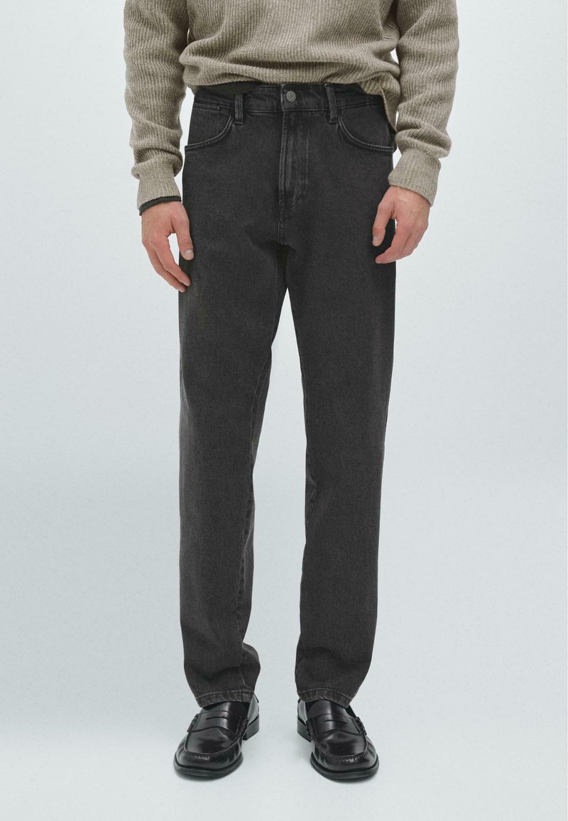 Ciemnoszare dżinsy w prostym kroju, pięć kieszeni i zapinane na guzik z przodu, w połączeniu z teksturowanym beżowym swetrem i czarnymi butami.